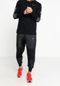 Schwarzes Langarmshirt mit Grafikdruck auf den Ärmeln, kombiniert mit schwarzen Panelhosen und roten Sportschuhen. Enthält Nike-Logos.