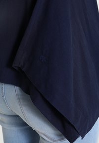 Camicia in cotone blu navy con orlo asimmetrico e dettaglio logo ricamato. Il tessuto sembra liscio con una texture leggera.