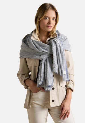 Blonde Frau, die eine beige Jacke und Hose trägt, mit einem großen blau-weiß gestreiften Schal, der ihr um die Schultern drapiert ist.