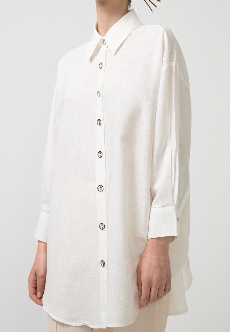 Personne portant une chemise blanche ample à manches longues, boutonnée, avec un col et de larges poignets. Seuls le haut du corps et la main sont visibles.