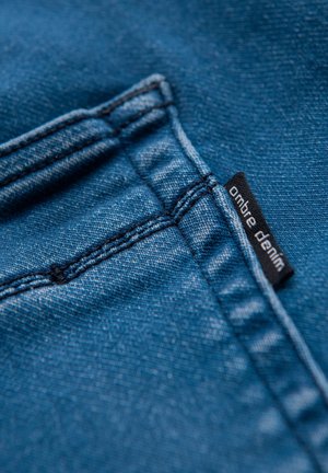 Niebieska tkanina dżinsowa o gładkiej fakturze, z kontrastowymi przeszyciami oraz czarną metką z napisem „ombre denim” umieszczoną przy kieszeni.