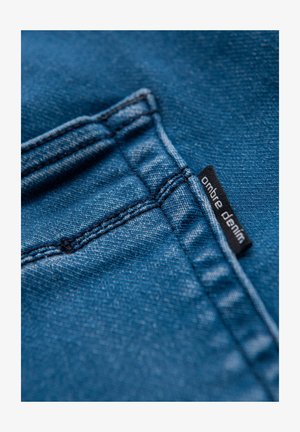 Blauwe denimstof met een gladde textuur, met contrasterende stikseldetails en een zwart stoffen label met de tekst "ombre denim" nabij de zak.