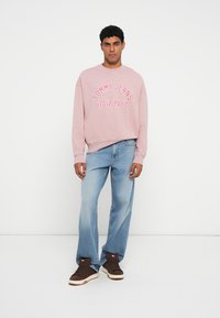 Gaiši rozā sporta krekls ar uzdrukātu "TOMMY JEANS NEW YORK CITY", saskaņots ar gaiši ziliem brīvā piegriezuma džinsiem un brūniem sporta apaviem.