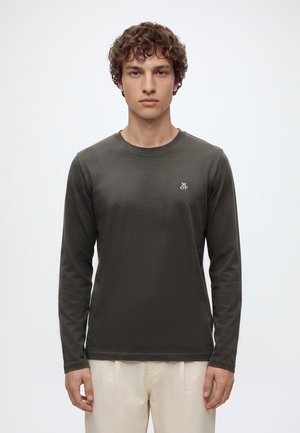 Jeune homme aux cheveux bouclés portant une chemise à manches longues vert foncé avec un petit logo blanc et un pantalon beige clair, debout devant un fond uni.