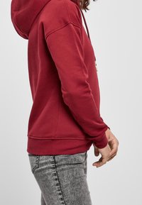 Sudadera roja con capucha, cordón ajustable y puños acanalados, hecha de tela suave. Se lleva con vaqueros grises, mostrando vista lateral y ajuste relajado.