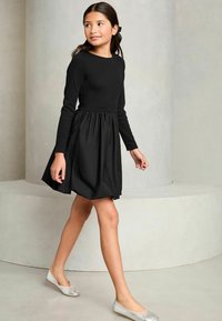 Schwarzes Kleid mit langen Ärmeln und einem gerafften Rock, kombiniert mit silbernen Ballerinas. Glatte Textur und schlichtes Design.