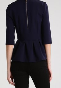 Námořnický modrý peplum top s přiléhavým korzetem a tříčtvrtečními rukávy; má kovový zip na zadní straně a hladkou, texturovanou tkaninu.
