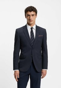 Giacca da abito blu navy, design strutturato con due bottoni, rever a punta e taschino sul petto con un accento a fantasia. Camicia bianca e cravatta.