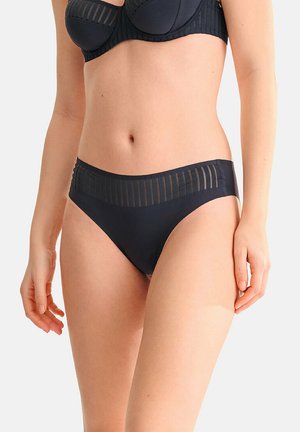 Femme portant un soutien-gorge noir et une culotte noire assortie avec des détails en rayures verticales transparentes, debout devant un fond clair uni.
