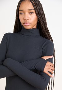 Blusa preta de mangas compridas com gola alta e textura canelada. Apresenta um pequeno logótipo em relevo no peito e mangas com buracos para os polegares.