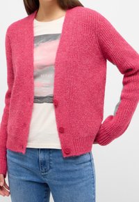 Rosa texturerad cardigan med långa ärmar, med V-ringning, två röda knappar, bärs över en ljusfärgad grafisk t-shirt.