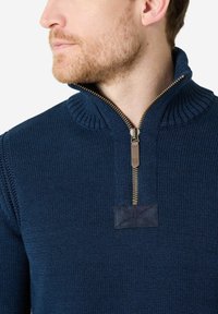 Marineblauer Strickpullover mit Reißverschlusskragen, geripptem Kragen und einem Lederpatch. Mit einem strukturierten Muster und einem Reißverschluss in Goldoptik.