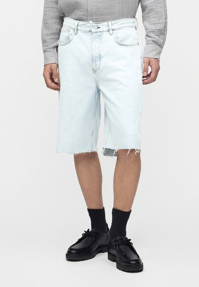 Homme portant un short en jean bleu clair effiloché, une chemise grise rayée à manches longues, des chaussettes noires et des chaussures en cuir noir à lacets.