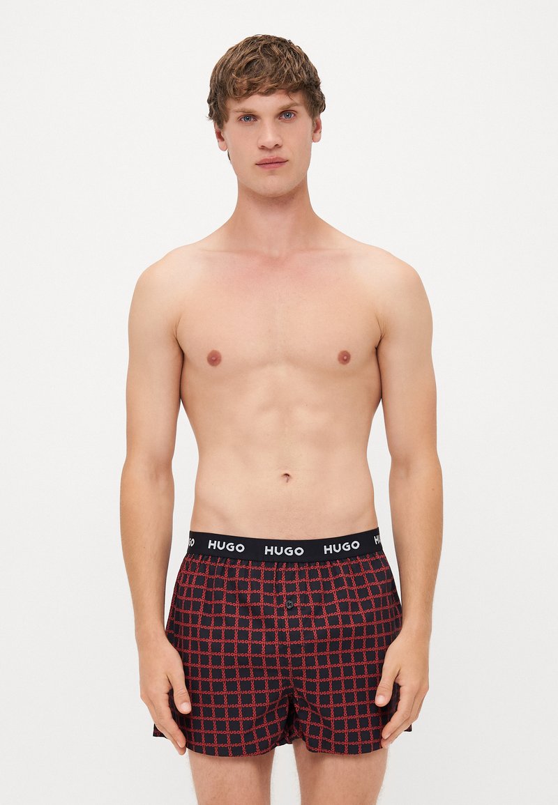 Boxers masculinos xadrez em vermelho e preto com um cós preto onde está o logotipo "HUGO", material em algodão, design até ao joelho.