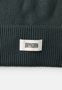 Zelena pletena kapa s rebrastom teksturom i bijelom etiketom na kojoj je ispisano "DRYKORN" crnim slovima.