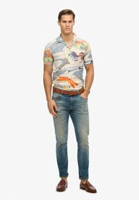 Kortarmpje, button-up shirt met een lichtblauwe basis, kleurrijke bloemen- en vogelpatronen; gecombineerd met lichtblauwe jeans en bruine instappers.