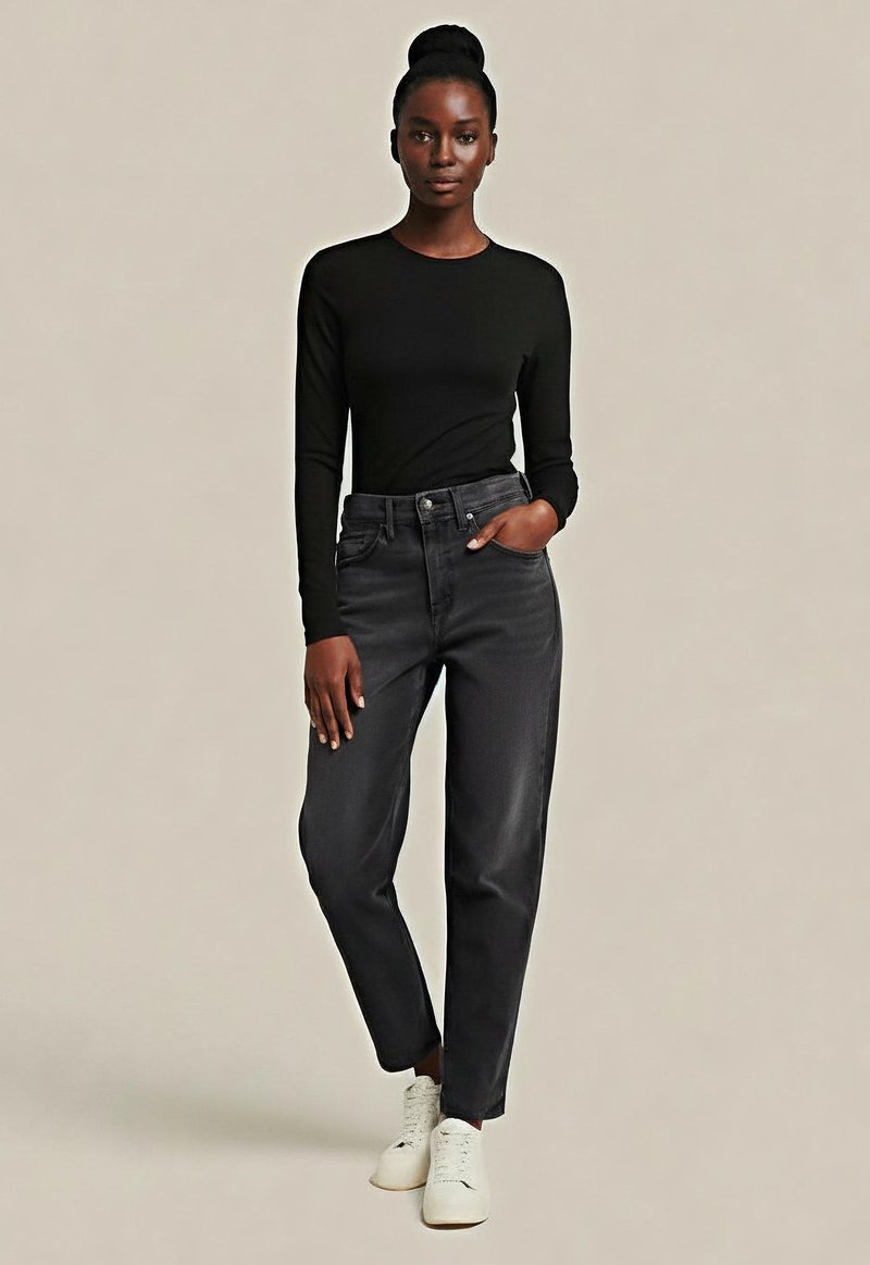 Kobieta o ciemnej skórze i włosach związanych w kok, ubrana w czarną koszulkę z długim rękawem, ciemnoszare dżinsy z wysokim stanem i białe trampki, stoi z jedną ręką w kieszeni.