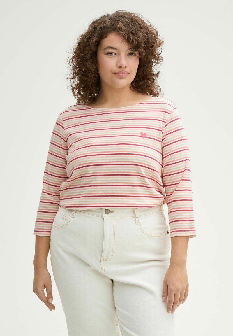 Camicia a maniche lunghe a righe di un rosa chiaro, con righe rosse e rosa scuro e un piccolo motivo a cuore sul petto. Abbinata a jeans bianchi.