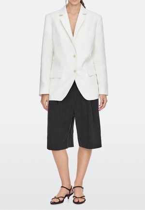 Femme portant un blazer blanc cintré, un short noir large mi-genoux et des sandales noires à talons, debout devant un fond uni.