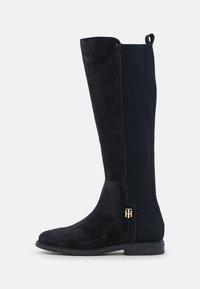 Botte en suede noire jusqu'au genou avec panneau élastique nervuré à l'arrière, semelle plate et petit logo doré "TH" près du talon.