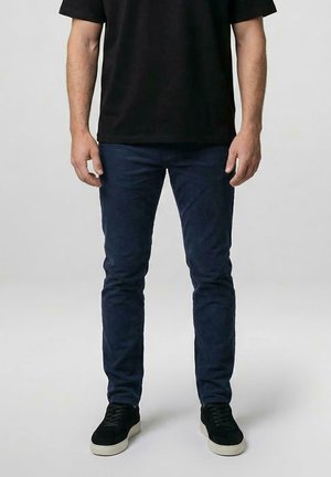 Man draagt donkerblauwe slim-fit broek, zwart T-shirt en zwarte sneakers met witte zolen, staand tegen een effen lichte achtergrond.