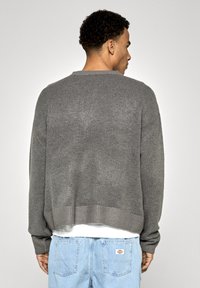 Pelēks adīts džemperis ar apaļu kakla izgriezumu un ribotām piedurknēm; tam ir brīvs piegriezums un mīksta tekstūra, apvienots ar gaiši ziliem džinsiem.