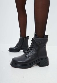 Bottines en cuir noir avec un laçage à l'avant, un bout arrondi et un talon carré avec un accent texturé. Détails de couture subtils visibles.