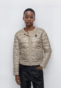 Beige vadderad jacka med rund halsringning, tryckknappar och två framfickor. Slät syntetisk material med en subtil lyster och strukturerad mönster.