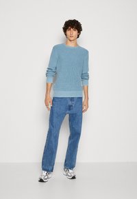 Helle blaue Strickjacke mit geripptem Saum und Bündchen, kombiniert mit blauen Jeans im geraden Schnitt und weißen Sneakers mit grauen Akzenten.