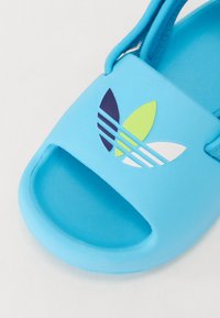 Sininen liukumäki-sandaali, jossa on teksturoitu päällinen, leikattu varvasosa ja monivärinen logo, jossa on vihreitä, valkoisia, tummansinisiä ja vaaleansinisiä yksityiskohtia.