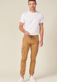 Tan cargobyxor med elastiska mudd, två sidofickor och dragsko i midjan, i kombination med en enkel vit t-shirt och vita sneakers.