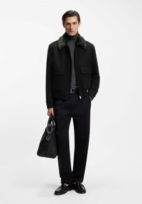 Mann in schwarzer Jacke mit Fellkragen, grauem Rollkragenpullover, schwarzer Hose, Slippern, hält schwarze Ledertasche und steht vor weißem Hintergrund.