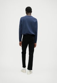 Temno modra pulover s dolgimi rokavi in krožnim izrezom, v kombinaciji s črnimi tesnimi jeans hlačami in belimi supergami. Pogledu zadaj.