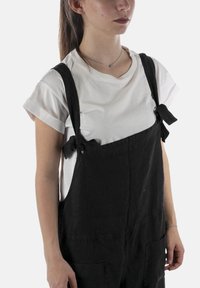 Salopette nera in lino con fiocchi regolabili sulle bretelle, indossata sopra una T-shirt bianca con maniche arrotolate. Presenta due tasche frontali.