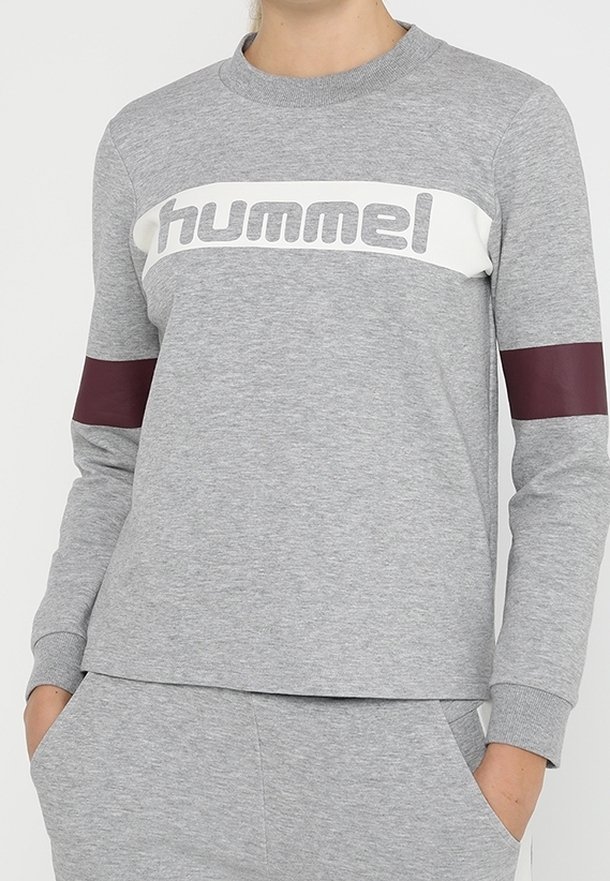 Personne portant un sweat-shirt gris avec le logo « hummel » sur la poitrine et des bandes bordeaux sur les deux manches, les mains dans les poches du pantalon gris assorti.