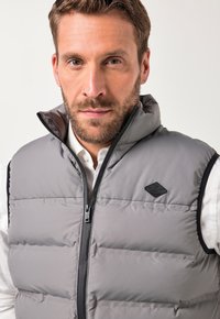 Graue Steppweste mit Reißverschluss, hoher Kragen, schwarzem Besatz und Logo-Patch. Strukturiert mit ausgeprägten gesteppten Abschnitten.