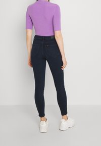 Jeans ajustados de mezclilla azul oscuro, con una cintura alta, cinco bolsillos y un diseño con ligero stretch. Combinados con una blusa corta morada y zapatillas blancas.