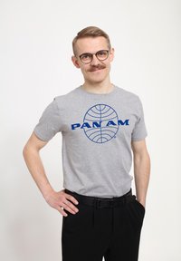 Šedé tričko s krátkým rukávem s modrým logem "PAN AM" a designem globusu na přední straně, v kombinaci s černými kalhotami a černým páskem.