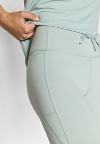 Ελαστικό κολάν σε ανοιχτό μέντα πράσινο χρώμα, κατασκευασμένο από ελαστικό υλικό με λεπτομερείς ραφές και ζωνάκι με κορδόνι στη μέση, παρουσιάζεται μαζί με ένα εφαρμοστό τοπ.