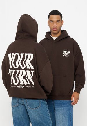 Deux jeunes hommes portant des sweat-shirts à capuche marron foncé, l’un faisant face vers l’avant et l’autre vers l’arrière, affichant "YOUR TURN" en grand texte blanc au dos.