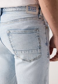 Jean en denim bleu clair avec une poche arrière ornée de coutures jaunes et bleues. Une étiquette de marque est visible à la taille.