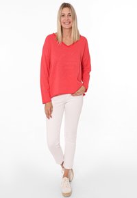 Maglione over-sized corallo con scollatura a V, caratterizzato da un messaggio positivo, abbinato a pantaloni skinny bianchi e scarpe stringate di colore chiaro.