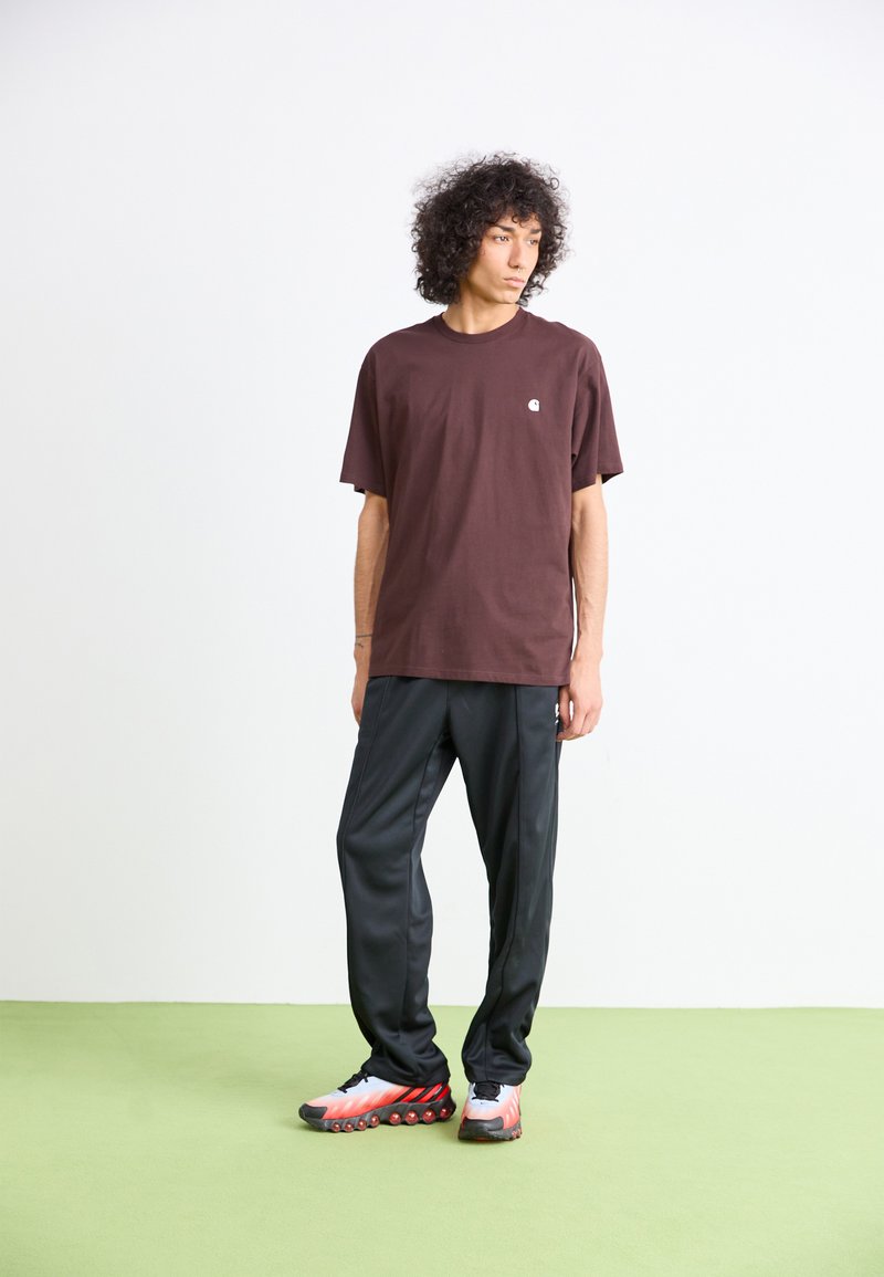 Giovane uomo con capelli ricci che indossa una maglietta marrone, pantaloni sportivi neri e sneakers bianco-rosse, in piedi su un pavimento verde chiaro contro un muro bianco.