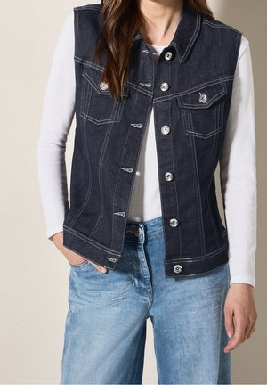 Vrouw draagt een donkerblauw denim gilet met knopen over een wit hemd met lange mouwen en lichtblauwe spijkerbroek, handen in de zakken, neutrale achtergrond.