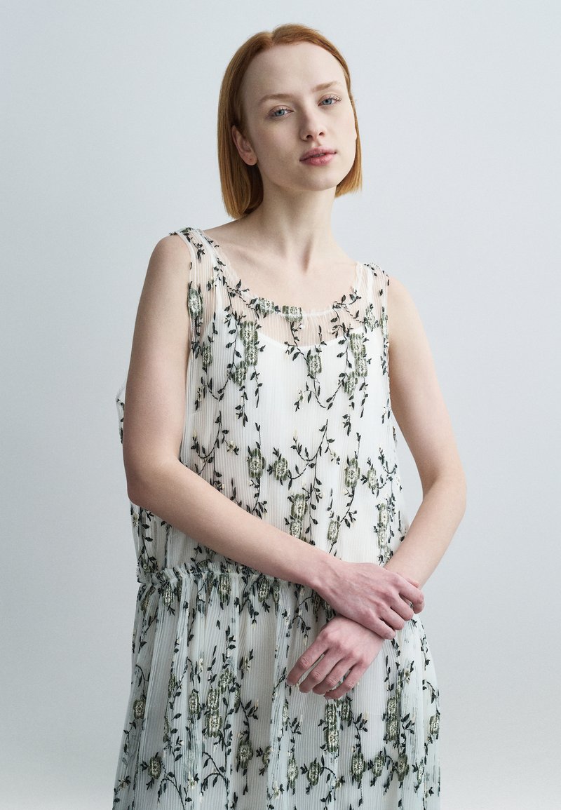 Robe sans manches en tissu blanc léger, ornée de broderies florales vertes et d'une texture plissée. Encolure ronde douce et taille froncée.