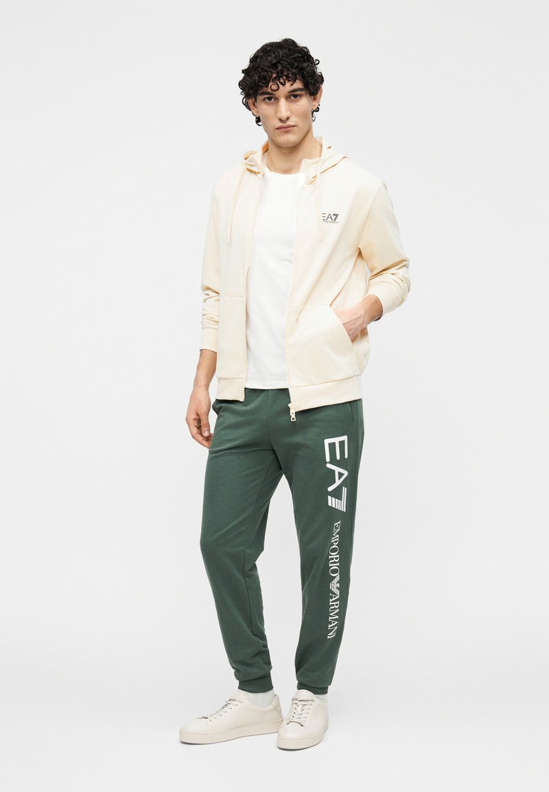 Tânăr cu păr creț, purtând un hanorac bej cu fermoar, tricou alb, pantaloni sport verzi cu textul „EA7 Emporio Armani” și adidași albi.