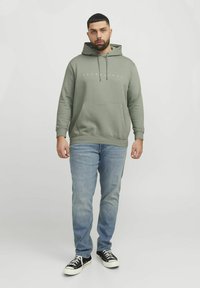 Grå hættetrøje med en lomme foran, lyseblå jeans og sorte sneakers. Hættetrøjen har et hvidt logo på brystet.