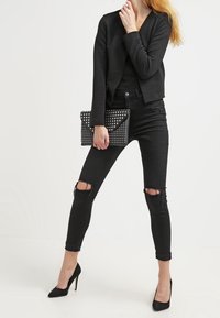 Czarna teksturowana kurtka, dopasowany czarny top, przetarte czarne skinny jeans oraz czarne szpilki. Trzyma w dłoni nabijany czarny kopertówkę.