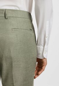 Jasnozielone spodnie z teksturowanej tkaniny o smukłym kroju, z tylną kieszenią i białą koszulą zapinaną na guziki, z widocznymi mankietami.