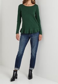 Green long-sleeve top with a flared hem, paired with blue jeans and black ankle boots. Smooth texture, round neckline, and minimal detailing.